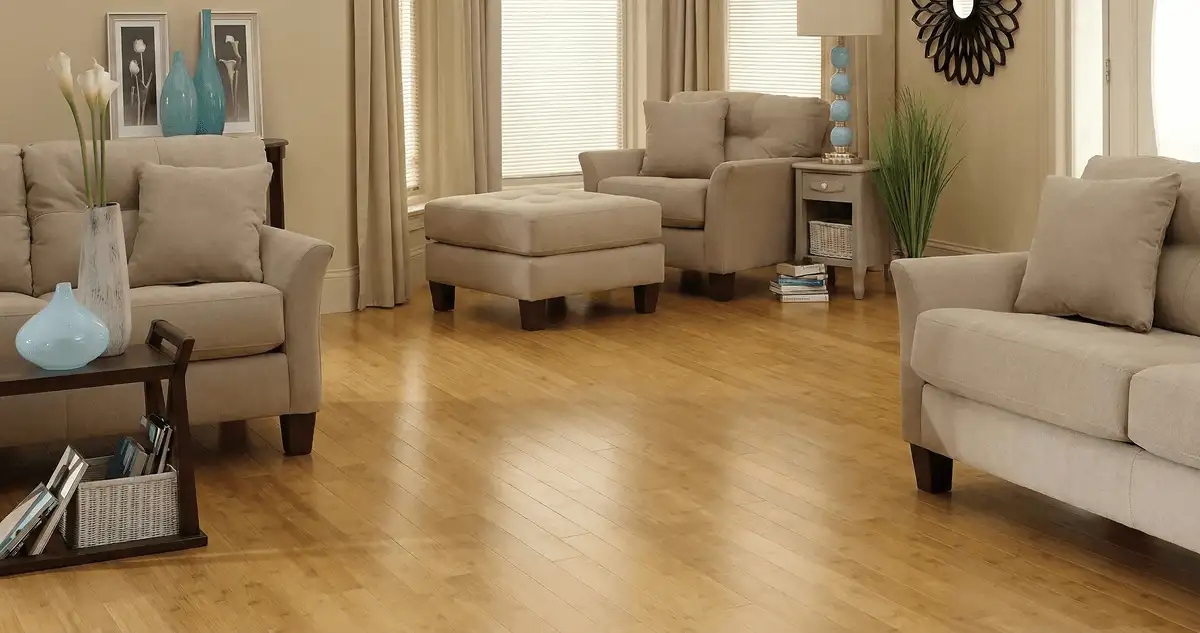 A living room with professionally installed bamboo flooring, beige sofas, a matching ottoman, a side table with a blue lamp, vases, framed pictures, and large windows with blinds and curtains in Marietta, GA